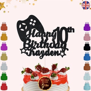 May include: Black glitter cake topper featuring a video game controller and the text "Happy 10th Birthday Kayden". The topper is displayed on a cake with red icing and strawberries.