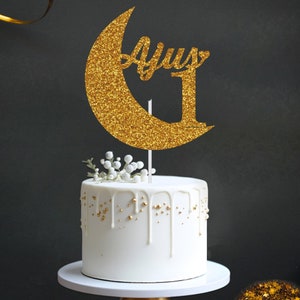 May include: Gold glitter moon cake topper with the word "Ayus" and the number "1" on it. The topper is on top of a white cake with gold drizzled icing.