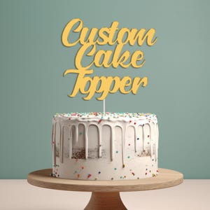 May include: A white cake with colourful sprinkles and white icing is topped with a gold wooden cake topper that says "Custom Cake Topper".