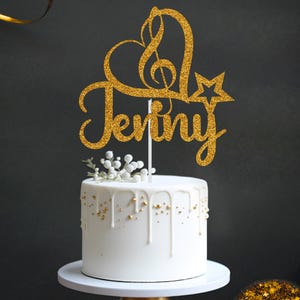 May include: Gold glitter cake topper featuring a heart, music note, and the name "Jenny". The topper is placed on a white cake with gold drizzled icing.