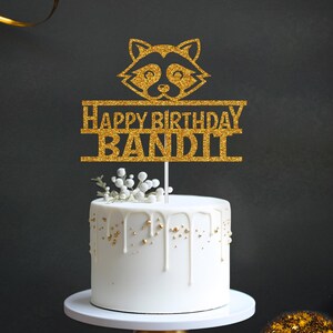 May include: Gold glitter cake topper with a raccoon silhouette and the text "Happy Birthday Bandit".