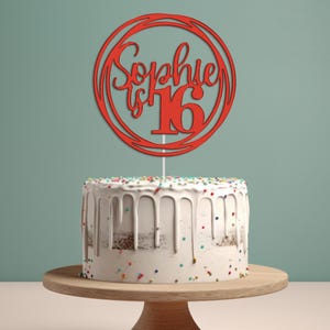 May include: Red cake topper with the text "Sophie is 16" in a circle design. The topper is on top of a white cake with colorful sprinkles.