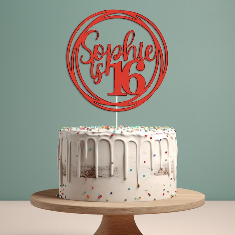 May include: Red cake topper with the text "Sophie is 16" in a circle design. The topper is on top of a white cake with colorful sprinkles.