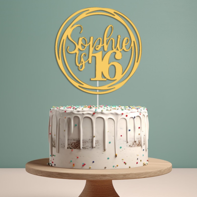 May include: Gold cake topper with the text "Sophie is 16" in a circle. The topper is on a white cake with colorful sprinkles.