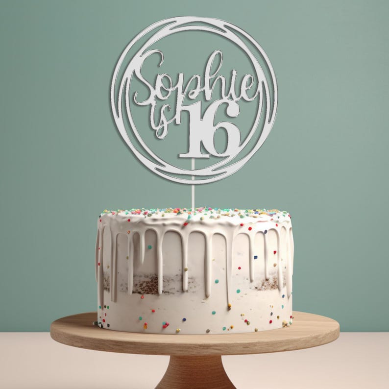 May include: White cake topper with a circle design and the text "Sophie is 16" in a cursive font. The topper is on top of a white cake with colorful sprinkles.