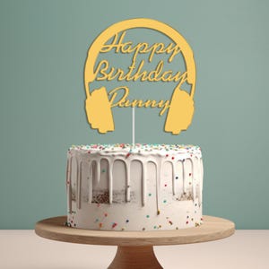 May include: A yellow cake topper shaped like headphones with the text "Happy Birthday Dummy" on it. The topper is on top of a white cake with colourful sprinkles.