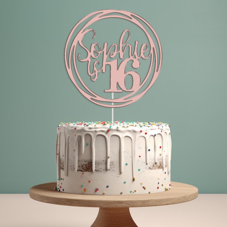 May include: Pink cake topper with the text "Sophie is 16" in a circle. The topper is on a white cake with colorful sprinkles.