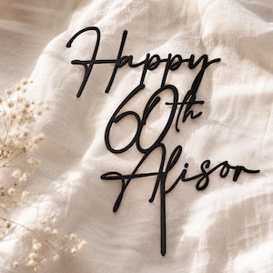 May include: Black cake topper with the words "Happy 60th Alison" in a cursive font. The topper is set against a white, textured background with delicate white flowers in the corner.