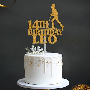May include: Gold glitter cake topper featuring a silhouette of a person on a scooter. The topper reads "14th Birthday Leo".