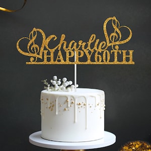 May include: Gold glitter cake topper with a treble clef and heart design. The topper says "Charlie Happy 60th".