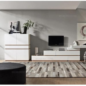 May include: A white storage cabinet with horizontal wood accents, a low-profile white media console with wood trim, and a matching white desk with a light-colored chair. A round black ottoman sits in the foreground on a gray patterned area rug.