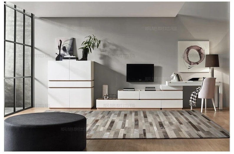 May include: A white storage cabinet with horizontal wood accents, a low-profile white media console with wood trim, and a matching white desk with a light-colored chair. A round black ottoman sits in the foreground on a gray patterned area rug.