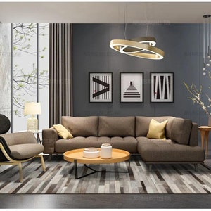 May include: A living room set featuring a brown sectional sofa, a matching armchair with a light wood frame, and a round light wood coffee table. The room includes a gray area rug, three framed black and white prints, and a modern gold ring pendant light.