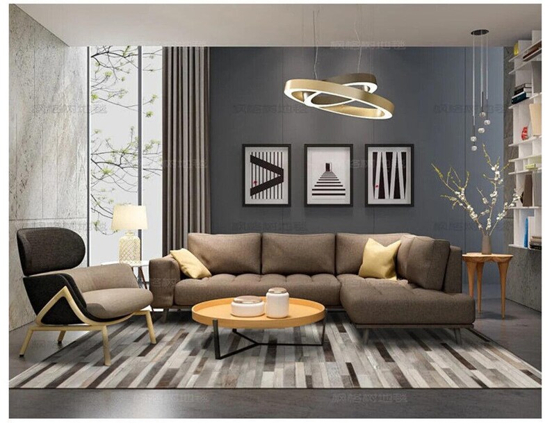 May include: A living room set featuring a brown sectional sofa, a matching armchair with a light wood frame, and a round light wood coffee table. The room includes a gray area rug, three framed black and white prints, and a modern gold ring pendant light.