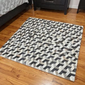Könnte beinhalten: Ein geometrisch gemusterter Teppich in Schwarz-, Grau- und Weißtönen. Der Teppich hat ein sich wiederholendes 3D-Design und ist rechteckig. Der Teppich liegt auf einem Parkettboden.