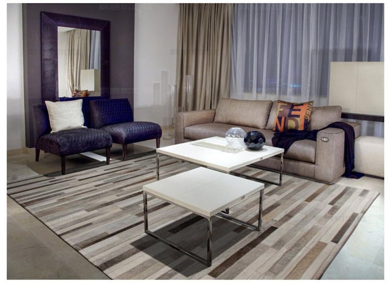 May include: A living room set featuring a beige sofa with a dark throw, two purple textured armchairs, and two square white coffee tables with chrome frames. A large rectangular area rug with a striped pattern in shades of brown, gray, and white.