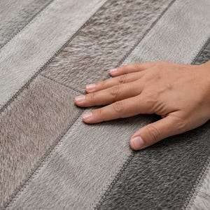 May include: A rug constructed from rectangular panels of genuine cowhide leather in varying shades of gray, charcoal, and silver. The panels are arranged in a linear pattern and joined together with visible, tight, tonal stitching throughout.