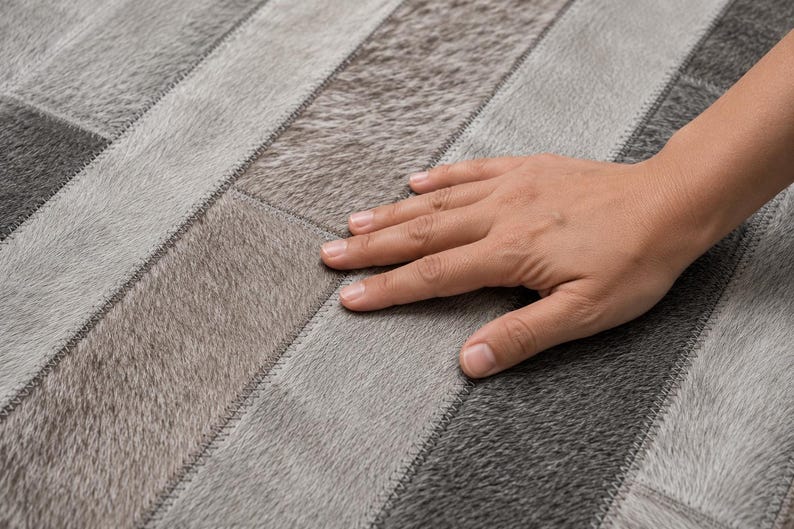 May include: A rug constructed from rectangular panels of genuine cowhide leather in varying shades of gray, charcoal, and silver. The panels are arranged in a linear pattern and joined together with visible, tight, tonal stitching throughout.