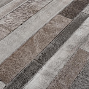 May include: A rectangular area rug constructed from stitched strips of natural cowhide leather. The surface features a patchwork pattern of varying textures and shades, including light gray, charcoal, and muted brown tones with visible stitching.