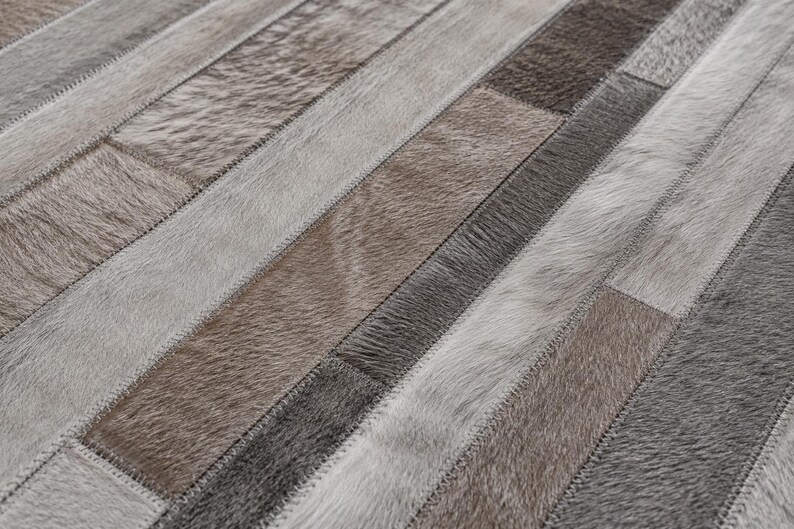 May include: A rectangular area rug constructed from stitched strips of natural cowhide leather. The surface features a patchwork pattern of varying textures and shades, including light gray, charcoal, and muted brown tones with visible stitching.