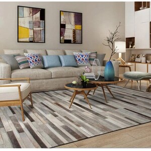 May include: A rectangular area rug featuring a multi-tonal pattern of horizontal gray, white, and brown rectangular strips. The rug is positioned in a living room setting with a beige sofa, a wooden armchair, and two round black coffee tables.