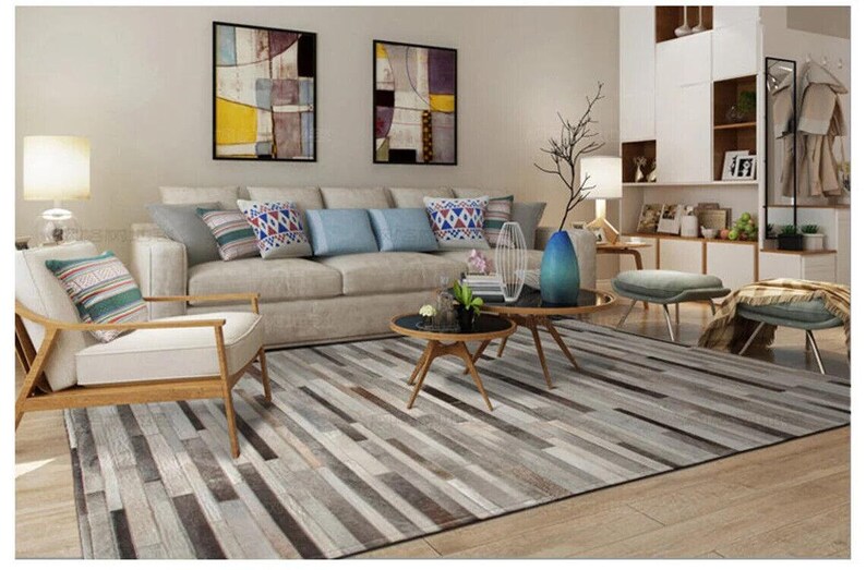 May include: A rectangular area rug featuring a multi-tonal pattern of horizontal gray, white, and brown rectangular strips. The rug is positioned in a living room setting with a beige sofa, a wooden armchair, and two round black coffee tables.