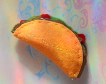 May include: A handmade felt taco with orange, green, brown, and red details. The taco is stitched with yellow thread and rests on a colorful, swirled background. A fun, whimsical food item.