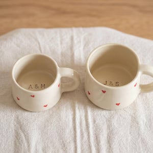 May include: Two off-white ceramic mugs with small red heart patterns on the exterior. The smaller mug features the text A & M printed on the interior base, while the larger mug features the text J & S printed on the interior base.