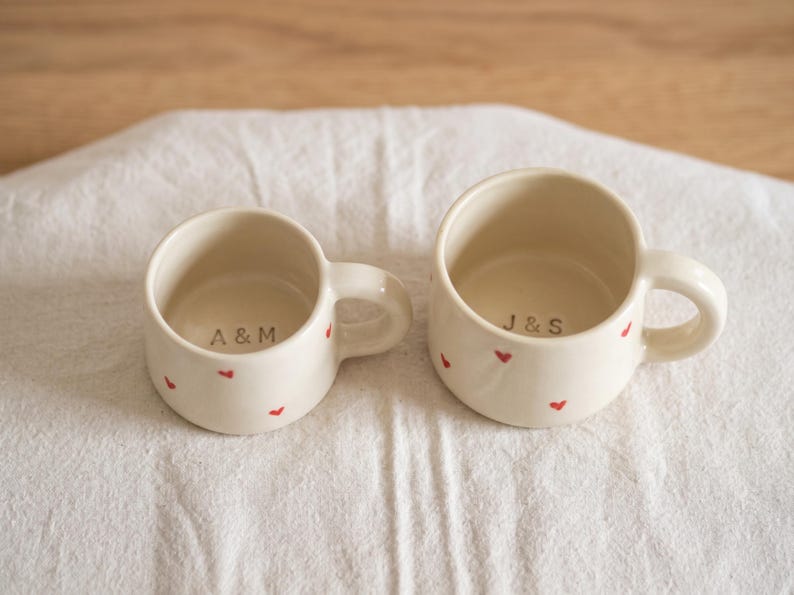 May include: Two off-white ceramic mugs with small red heart patterns on the exterior. The smaller mug features the text A & M printed on the interior base, while the larger mug features the text J & S printed on the interior base.