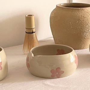 Pu&ograve; includere: Una collezione di oggetti in ceramica che comprende due ciotole color crema con fiori rosa dipinti a mano, un frustino per matcha in bamb&ugrave;, un grande vaso in argilla beige testurizzato e un vaso liscio bianco sporco su una superficie bianca.