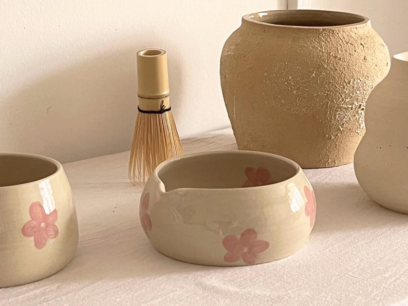 Pu&ograve; includere: Una collezione di oggetti in ceramica che comprende due ciotole color crema con fiori rosa dipinti a mano, un frustino per matcha in bamb&ugrave;, un grande vaso in argilla beige testurizzato e un vaso liscio bianco sporco su una superficie bianca.