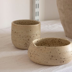 May include: Two speckled ceramic bowls in a neutral beige color. One is a small, rounded bowl with a wide opening, and the other is a taller, cylindrical cup. Both have a textured surface and dark speckles.