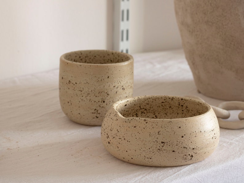 May include: Two speckled ceramic bowls in a neutral beige color. One is a small, rounded bowl with a wide opening, and the other is a taller, cylindrical cup. Both have a textured surface and dark speckles.