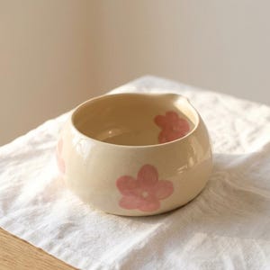 Pu&ograve; includere: Una ciotola in ceramica color crema di forma rotonda, caratterizzata da una finitura liscia e lucida e da motivi floreali rosa dipinti a mano. La ciotola presenta un'apertura ampia e un piccolo beccuccio discreto su un lato.