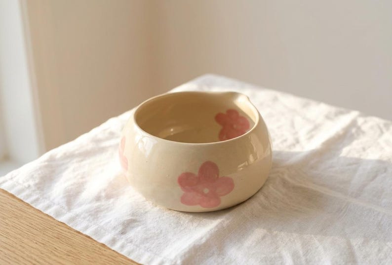 Pu&ograve; includere: Una ciotola in ceramica color crema di forma rotonda, caratterizzata da una finitura liscia e lucida e da motivi floreali rosa dipinti a mano. La ciotola presenta un'apertura ampia e un piccolo beccuccio discreto su un lato.