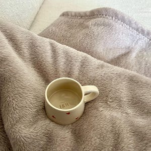 May include: A small, cream-colored ceramic mug with a single handle, featuring tiny red heart patterns on the exterior and the letters "I&H" printed in a dark color on the interior base, resting on a soft, textured, light brown plush fabric.