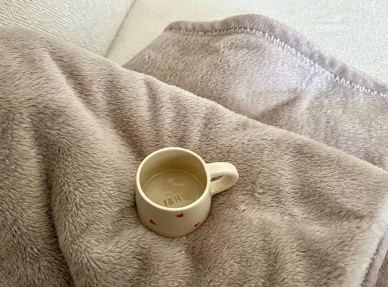 May include: A small, cream-colored ceramic mug with a single handle, featuring tiny red heart patterns on the exterior and the letters "I&H" printed in a dark color on the interior base, resting on a soft, textured, light brown plush fabric.