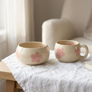 Pu&ograve; includere: Una ciotola in ceramica e una tazza abbinata, entrambe con base color crema e motivi floreali rosa dipinti a mano. La tazza ha un corpo arrotondato e un manico curvo, mentre la ciotola presenta un'apertura ampia e una finitura liscia.