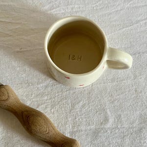May include: A cream-colored ceramic mug with small red heart patterns on the exterior and the letters I&H stamped into the interior base, positioned next to a metal pottery tool with a light brown wooden handle.