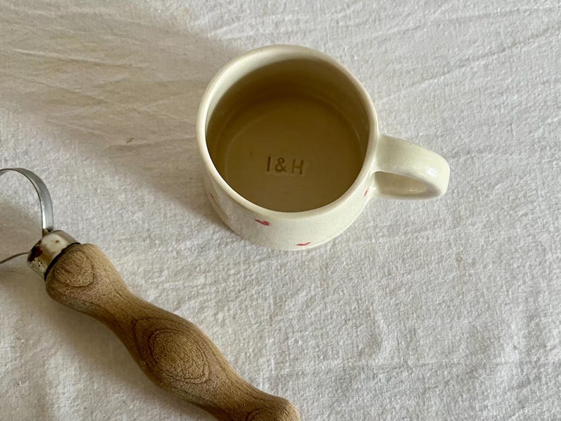 May include: A cream-colored ceramic mug with small red heart patterns on the exterior and the letters I&H stamped into the interior base, positioned next to a metal pottery tool with a light brown wooden handle.