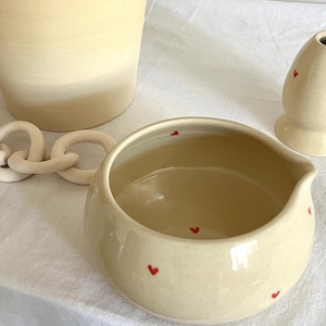 May include: Cream-colored ceramic pitcher, small bowl, and vase with red heart accents. A ceramic chain is next to the pitcher. The objects are on a white surface. The pitcher has a wide base and a narrow top.