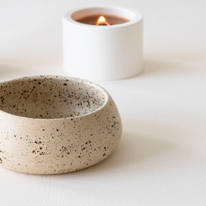 May include: A speckled, beige ceramic bowl with a rounded shape and a small spout. A white candle in a cylindrical holder with a lit flame is in the background. The items are on a white surface.