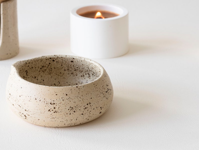 May include: A speckled, beige ceramic bowl with a rounded shape and a small spout. A white candle in a cylindrical holder with a lit flame is in the background. The items are on a white surface.