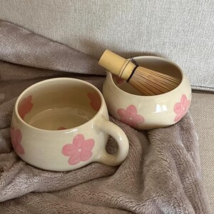 Pu&ograve; includere: Una tazza in ceramica color crema dalla forma arrotondata con manico, decorata con fiori rosa dipinti a mano. Accanto si trova una ciotola in ceramica abbinata color crema contenente un frustino per matcha in bamb&ugrave; marrone chiaro.