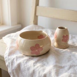 Pu&ograve; includere: Una ciotola in ceramica e un piccolo vaso abbinato, entrambi caratterizzati da uno smalto color crema maculato decorato con motivi floreali rosa dipinti a mano. La ciotola ha una forma arrotondata con un piccolo beccuccio, il vaso ha il collo stretto.