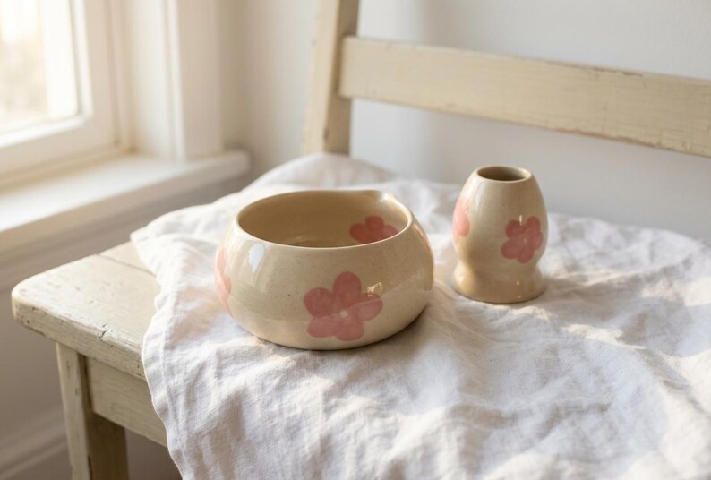 Pu&ograve; includere: Una ciotola in ceramica e un piccolo vaso abbinato, entrambi caratterizzati da uno smalto color crema maculato decorato con motivi floreali rosa dipinti a mano. La ciotola ha una forma arrotondata con un piccolo beccuccio, il vaso ha il collo stretto.