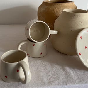 May include: A collection of cream-colored ceramic mugs and a lidded jar, each decorated with small red heart motifs. The mugs feature sturdy handles, and one mug interior is stamped with the letters I&H. A larger, textured tan vessel sits behind.