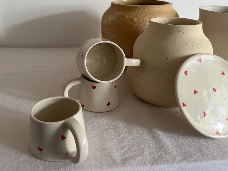 May include: A collection of cream-colored ceramic mugs and a lidded jar, each decorated with small red heart motifs. The mugs feature sturdy handles, and one mug interior is stamped with the letters I&H. A larger, textured tan vessel sits behind.