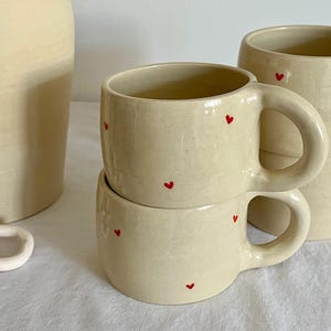 Puede incluir: Tazas de cerámica color crema con detalles de corazones rojos. Las tazas tienen forma redondeada con asa y están apiladas. En el fondo se ve un jarrón a juego. Las tazas están sobre una superficie blanca.