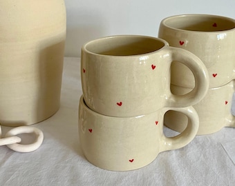 May include: Cream-colored ceramic mugs with red heart accents. The mugs have a rounded shape with a handle and are stacked. A matching vase is visible in the background. The mugs are on a white surface.